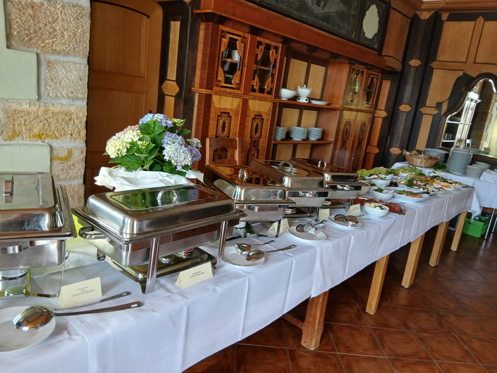 Buffet mit Metallwarmhaltern, weißen Tischdecken und Blumenarrangement im Holzraum