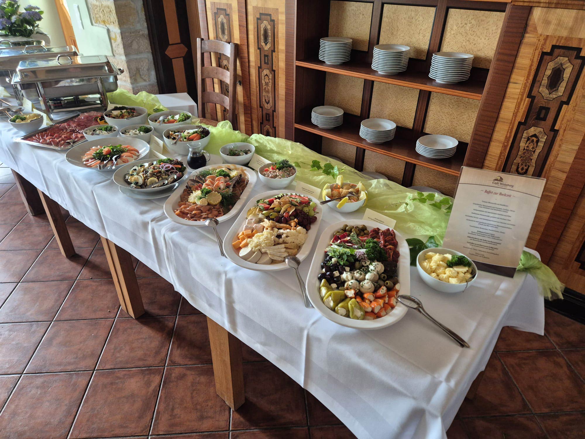 Buffet mit weißen Platten, bunten Salaten, Fleisch und Obst, Schild am Rand