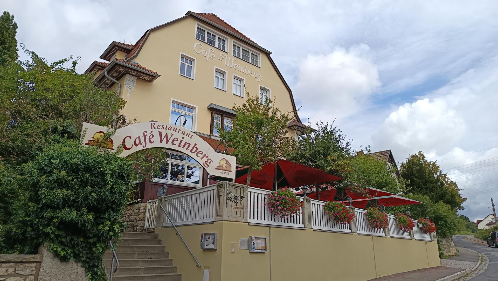 Gelbes Cafe mit roten Schirmen und Blumenkaesten am Hang, ruhig