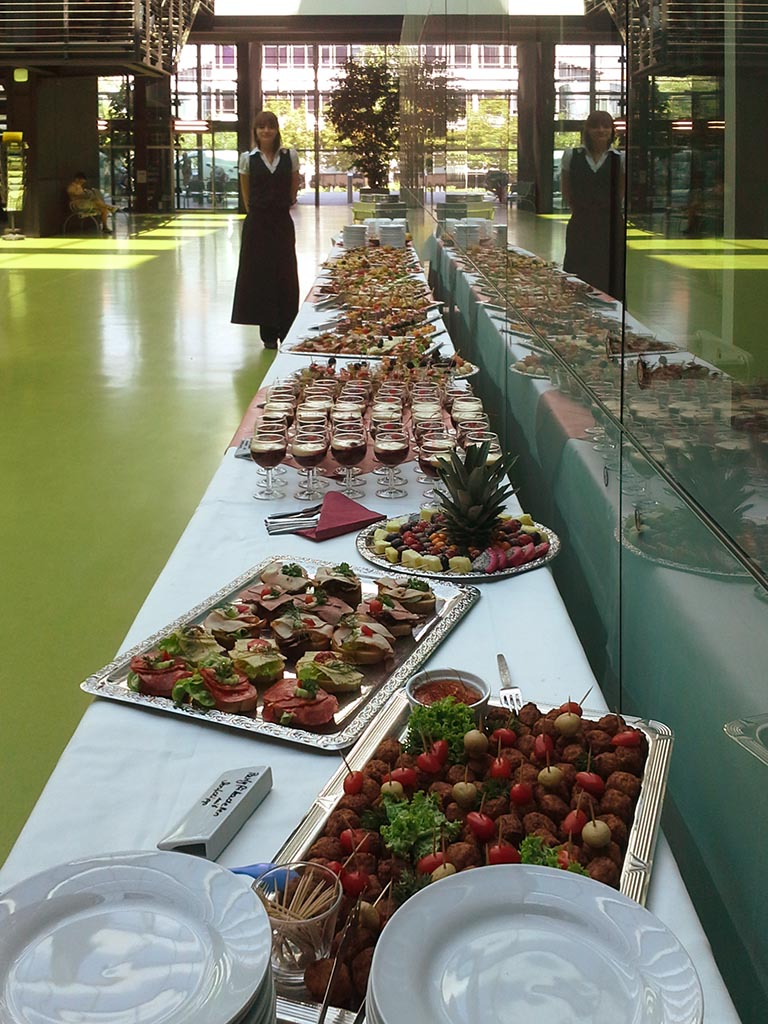 Zwei Kellner mit neutralem Blick bei langem Buffet mit Häppchen und Rotwein