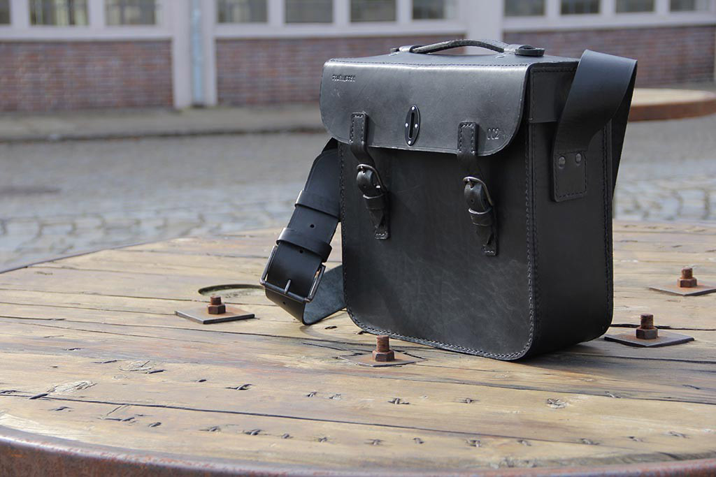 Schwarze Ledertasche mit ruhigem Ausdruck und breitem Schultergurt auf Holzspule