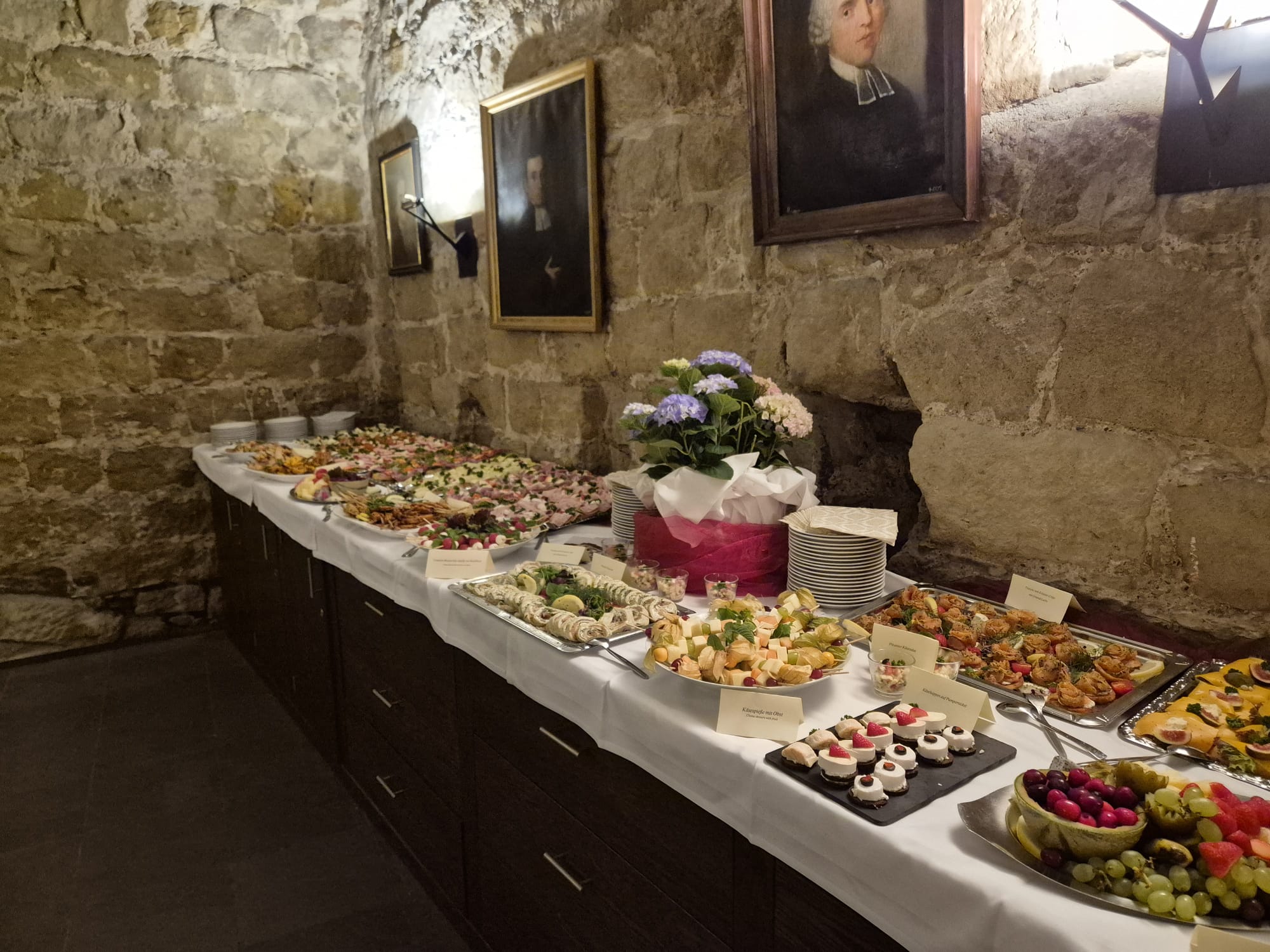 Buffet mit bunten Häppchen auf weißer Decke im Steingewölbe, Porträts an Wand